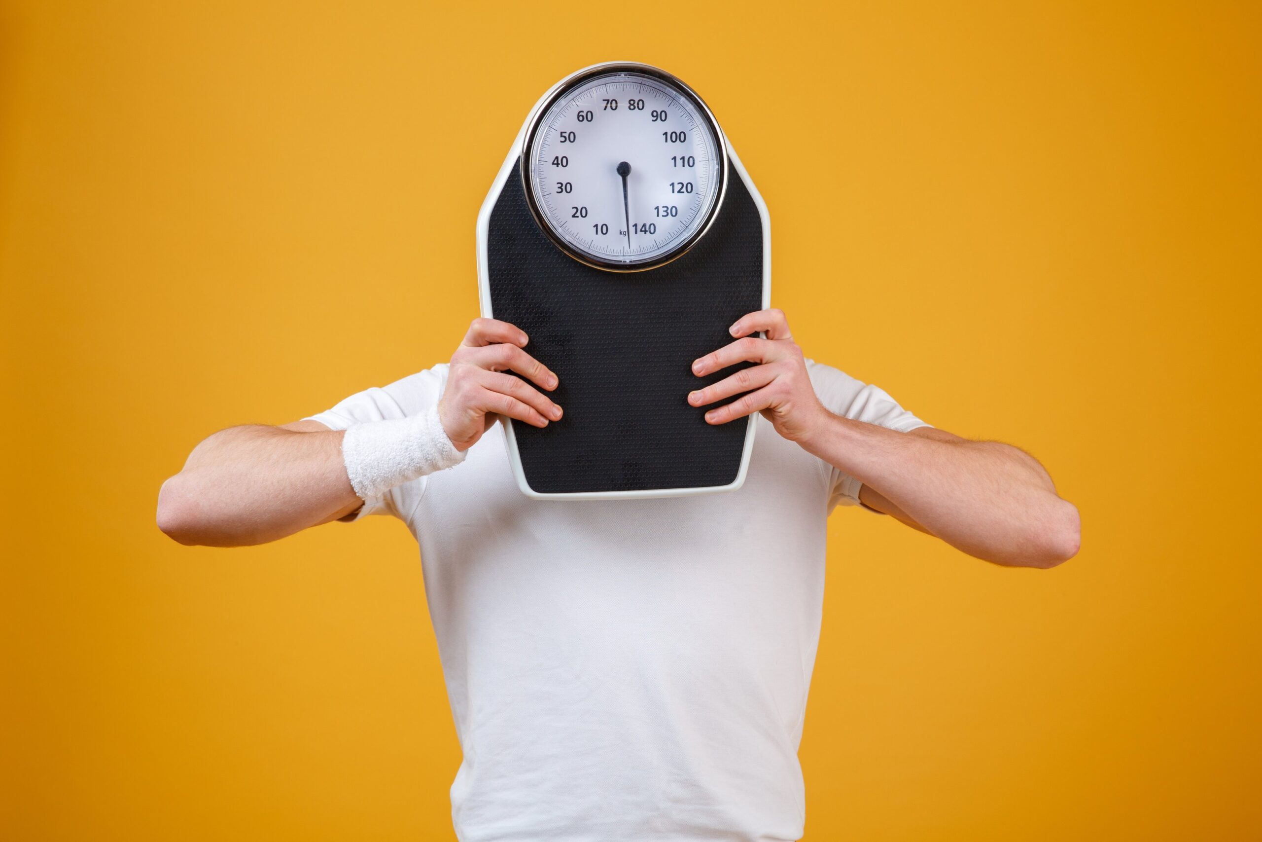 Boy hides his face behind weight measuring balance standing infront of mustard background.